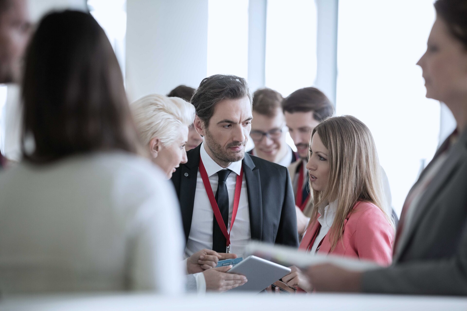 A man standing in a crowded room listening to two women, one on each side of him as they look at a document together. A man standing in a crowded room listening to two women, one on each side of him as they look at a document together.