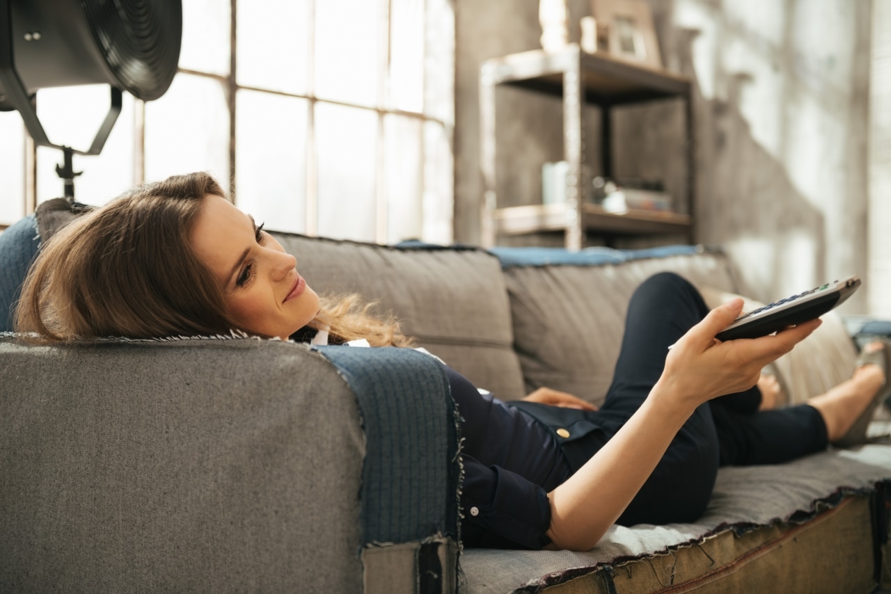 Mulher jovem morena relaxando em um sofá e assistindo à TV em um apartamento. Detalhes de decoração urbana e elegante, e janela.