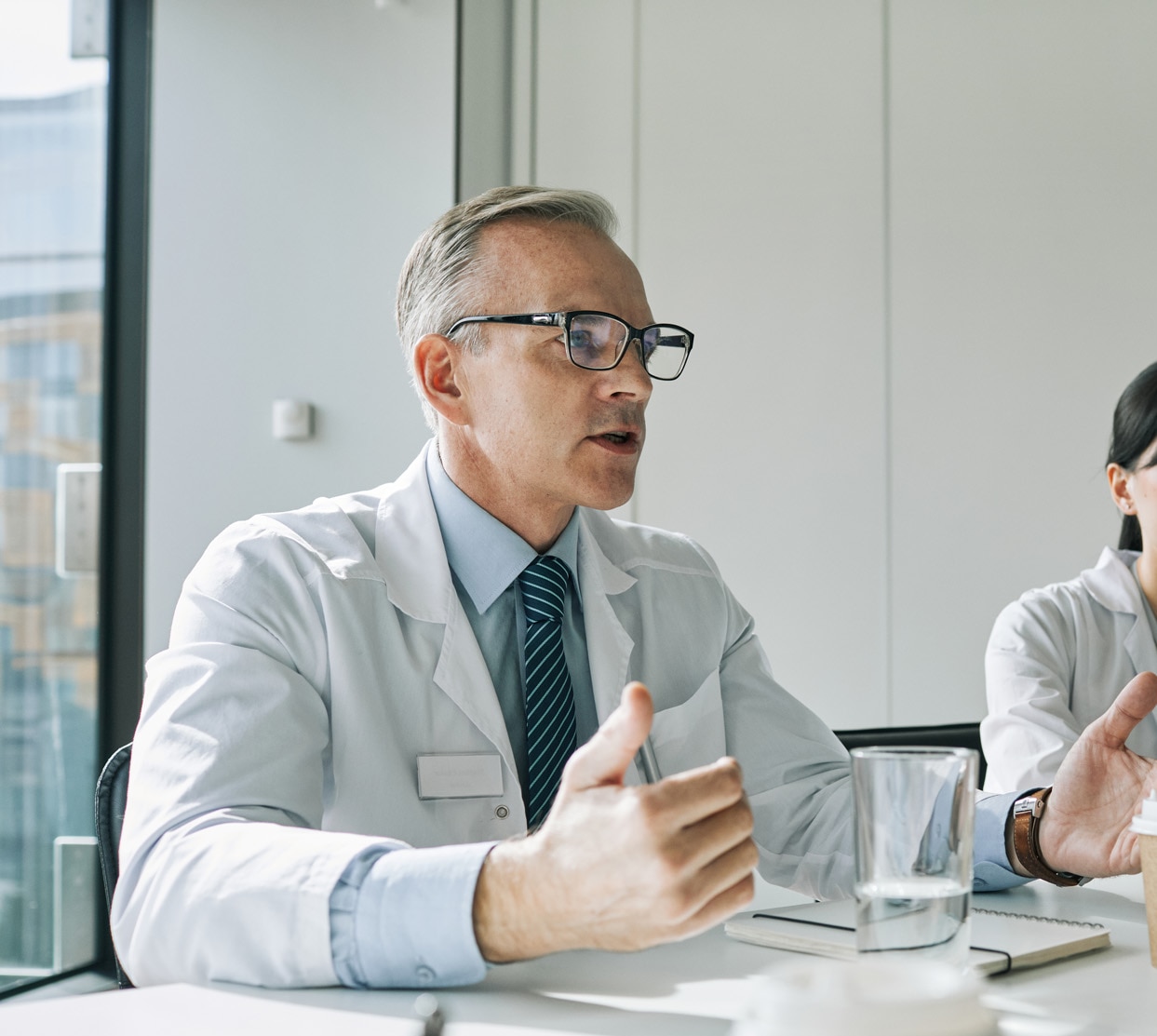 Portrait of mature doctor sitting at meeting table in conference room while speaking during medical seminar; Shutterstock ID 1987128383; purchase_order: -; job: -; client: -; other: -