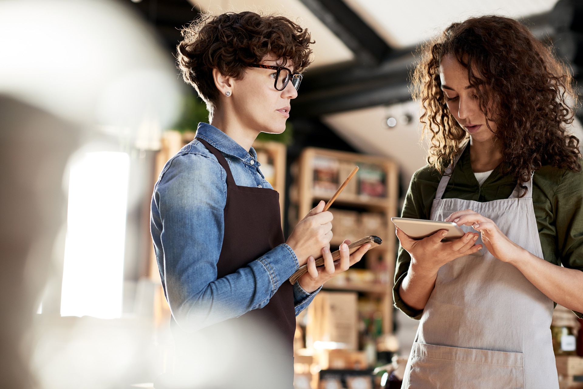 Running small business. Two curly concentrated female entrepreneurs in aprons working in their store