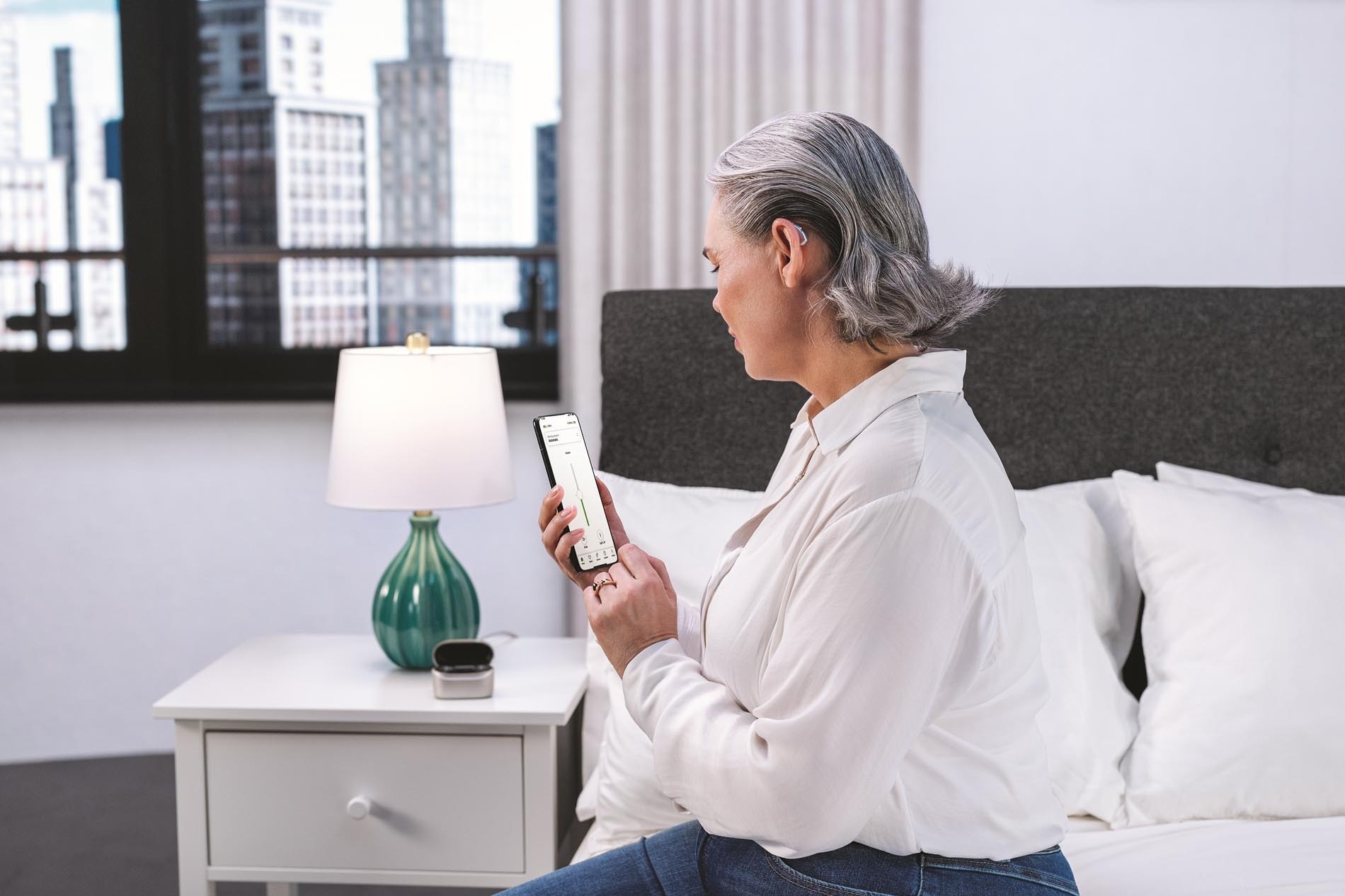 An elderly woman sitting on a bed in a brightly lit bedroom looking at her smartphone Eine ältere Frau sitzt auf einem Bett in einem hell beleuchteten Zimmer und schaut auf Ihr Smartphone